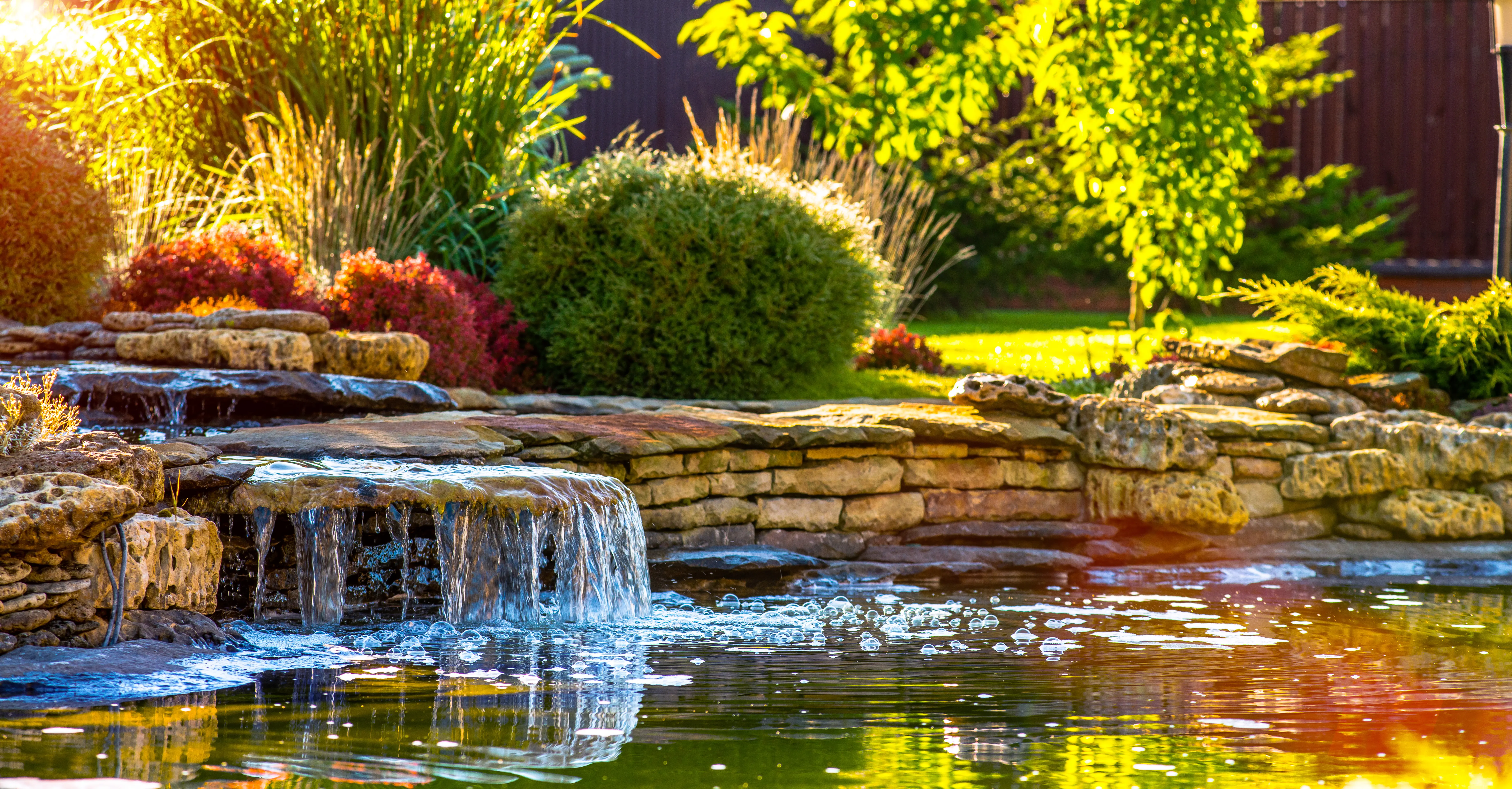 Gartenteich mit kleiner Naturstein-Wasserfall, klarer Wasseroberfläche, Seerand aus geschichteten Steinen und sonniger Bepflanzung im Hintergrund